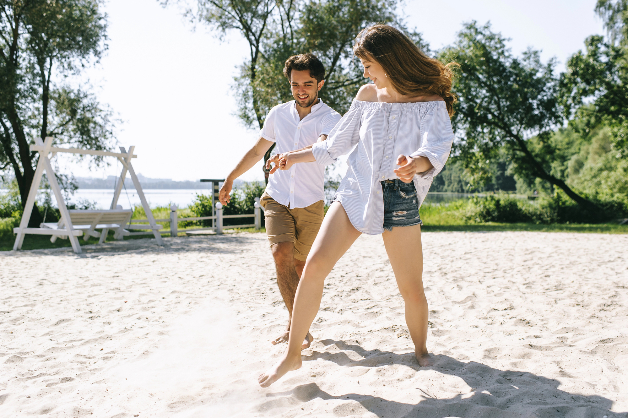 A young man and woman are joyfully running barefoot on a sandy beach. The man is wearing a white shirt and brown shorts, while the woman is in a white off-the-shoulder blouse and denim shorts. Trees and a swing are visible in the background on a sunny day.