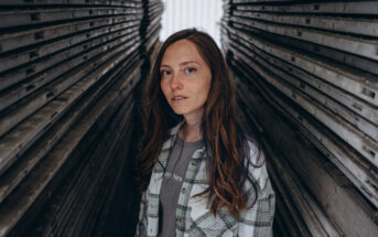 A young woman with long brown hair and freckles stands between two closely stacked rows of wooden planks. She is wearing a green and white plaid shirt over a gray T-shirt. She gazes directly at the camera with a neutral expression.