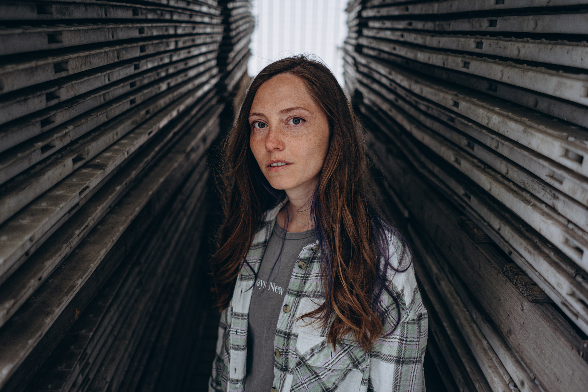 A young woman with long brown hair and freckles stands between two closely stacked rows of wooden planks. She is wearing a green and white plaid shirt over a gray T-shirt. She gazes directly at the camera with a neutral expression.