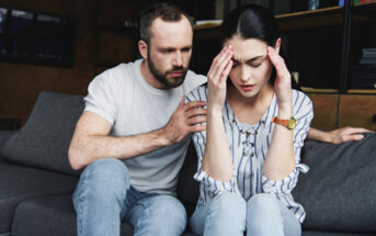 A man with a beard and wearing a white t-shirt sits on a gray couch, gently comforting a woman who looks distressed. She has her hands on her temples, appearing upset, and is wearing a striped blouse and a watch. Shelves with books are visible in the background.