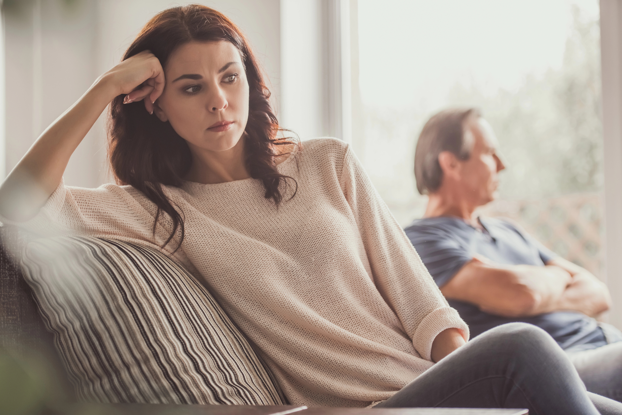 A woman with a worried expression sits on a couch resting her head on her hand. In the background, a man sits with his arms crossed and looks away. Both appear to be in a state of tension or disagreement. The setting is a bright room with natural light coming through the window.