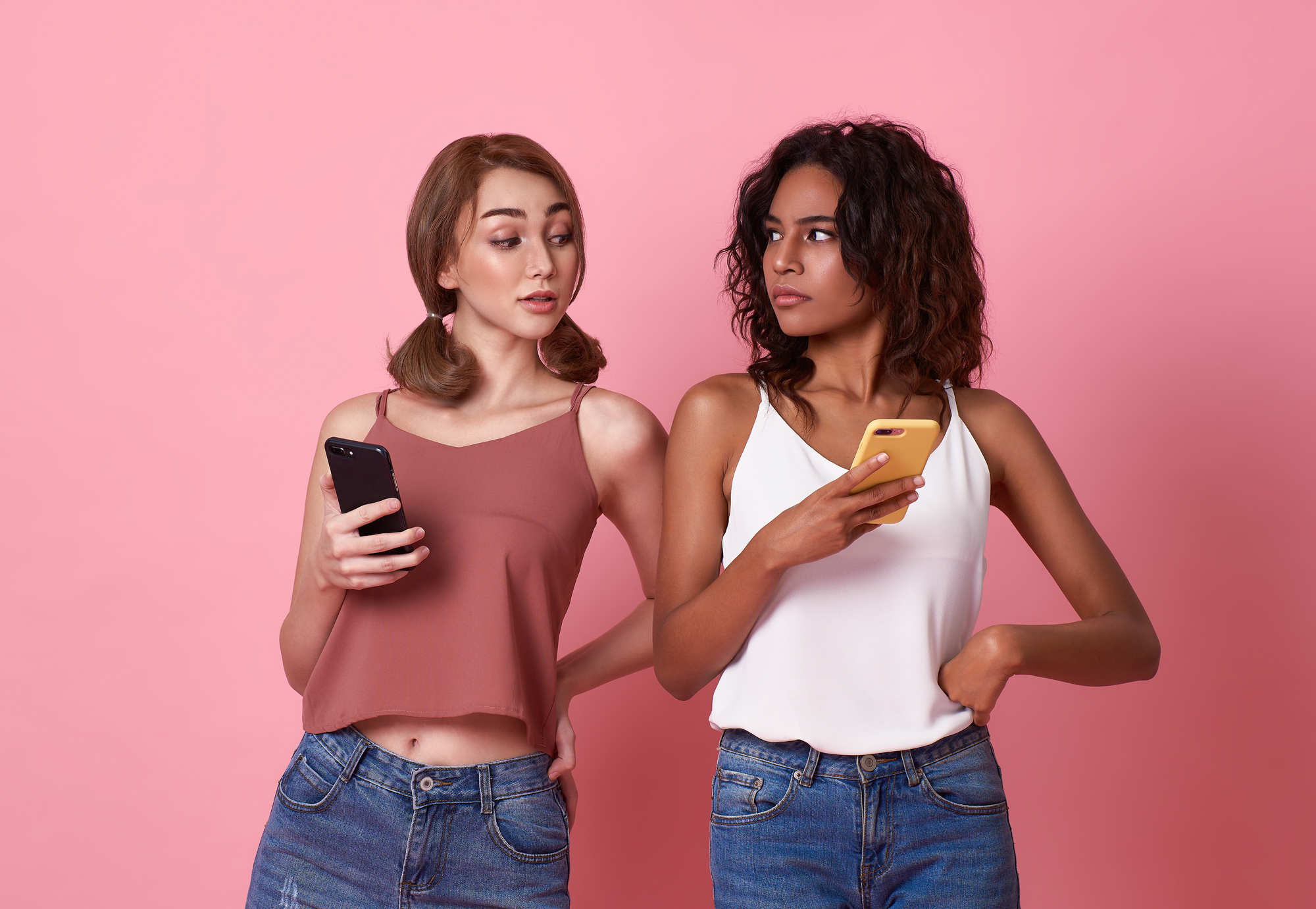 Two women standing against a pink background, looking at each other with raised eyebrows. Both are holding smartphones, one with a yellow case and the other with a black case. They are dressed casually in sleeveless tops and jeans, indicating a possible disagreement.