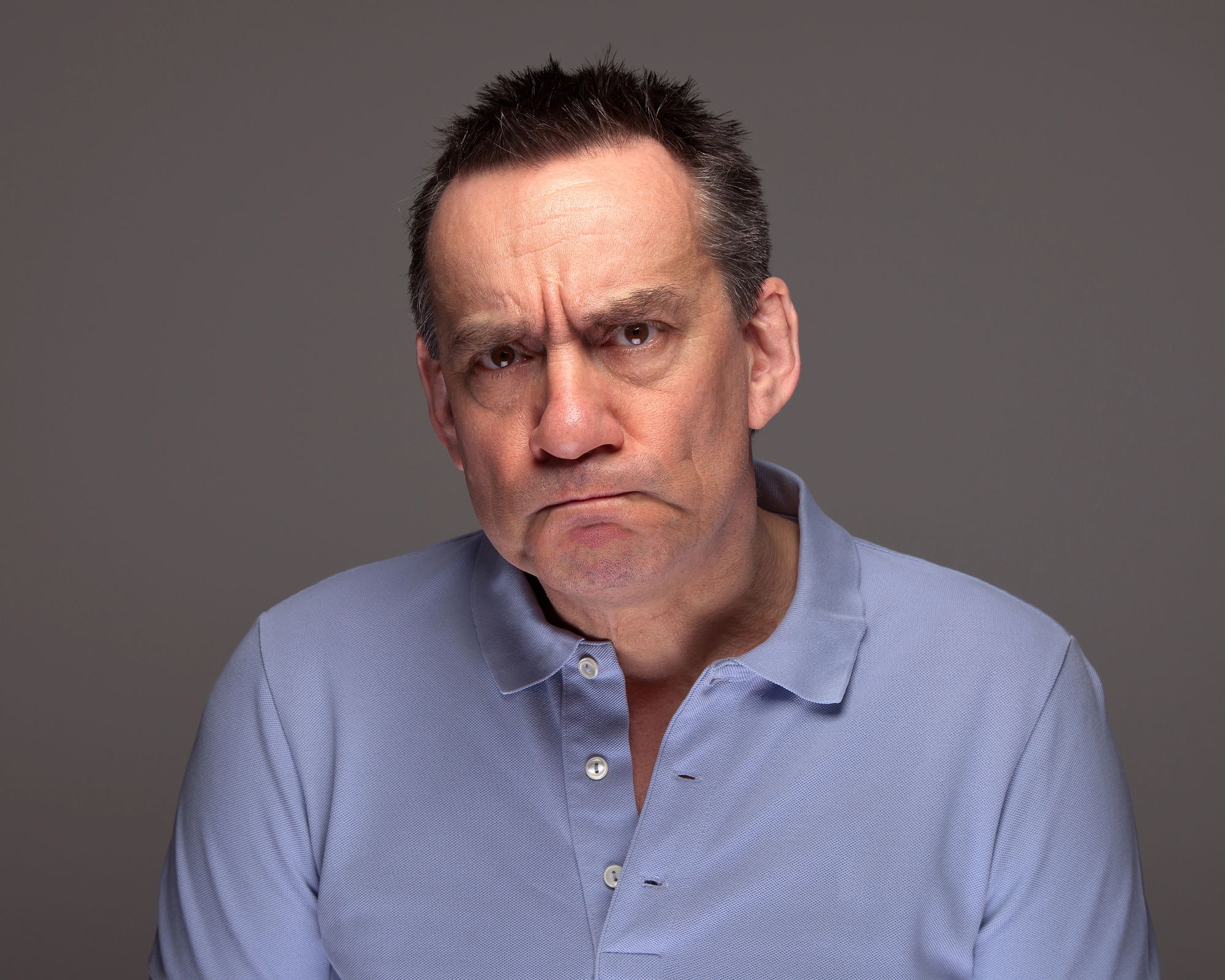 A middle-aged man with short dark hair and expressive eyebrows is wearing a light blue polo shirt. He has a serious and slightly furrowed expression on his face. The background is plain and gray.