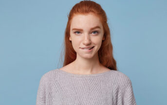 Young woman with long red hair, wearing a light gray sweater, stands against a solid blue background. She is looking at the camera with a slight, relaxed smile and has a light facial expression.