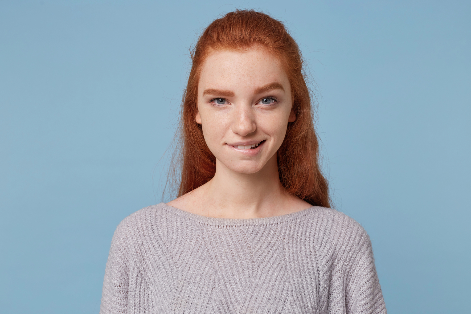 Young woman with long red hair, wearing a light gray sweater, stands against a solid blue background. She is looking at the camera with a slight, relaxed smile and has a light facial expression.