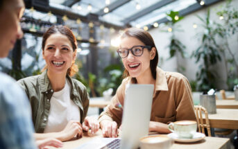 Three people are sitting in a well-lit café with greenery in the background, engaged in conversation and looking at a laptop screen. They are smiling and appear to be enjoying their time together. Two cups of coffee are on the table.