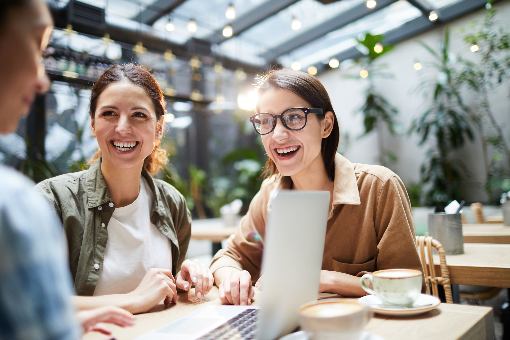 Three people are sitting in a well-lit café with greenery in the background, engaged in conversation and looking at a laptop screen. They are smiling and appear to be enjoying their time together. Two cups of coffee are on the table.