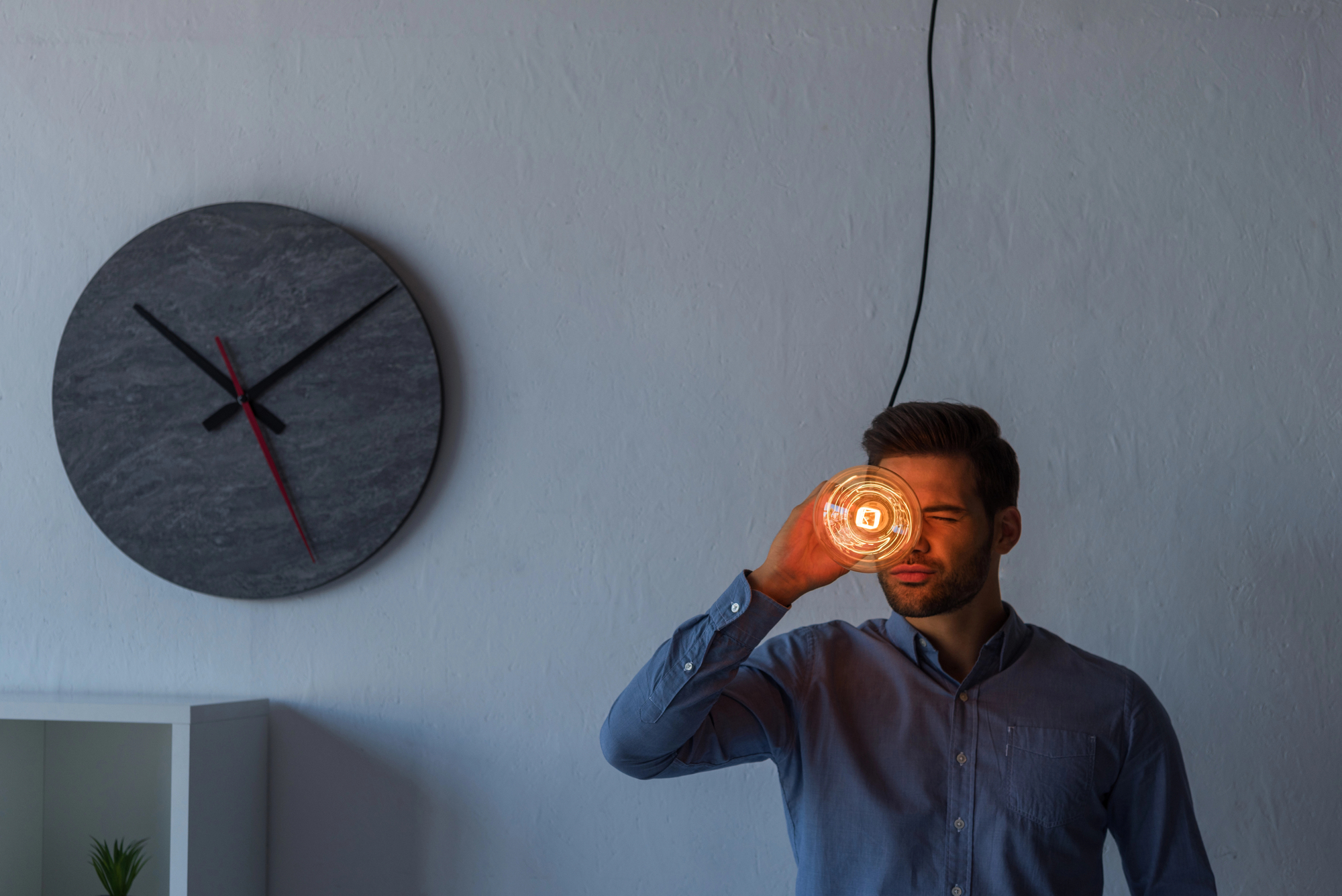 A man in a blue shirt is standing indoors, holding a lit light bulb close to his face and squinting. Behind him on the wall is a large circular clock with black and red hands, and below it is a small shelf with a plant.