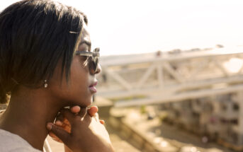 A person wearing sunglasses and a white top is shown in profile, resting their chin on their hands. They are outside, with blurred background elements of an industrial, urban setting featuring white bridge structures and buildings.