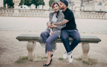 A couple sits on a stone bench outdoors. The man, wearing a cap and dark clothing, embraces the woman from behind, smiling. The woman, wearing a patterned jacket and purple pants, closes her eyes and smiles. The background features historical architecture and statues.