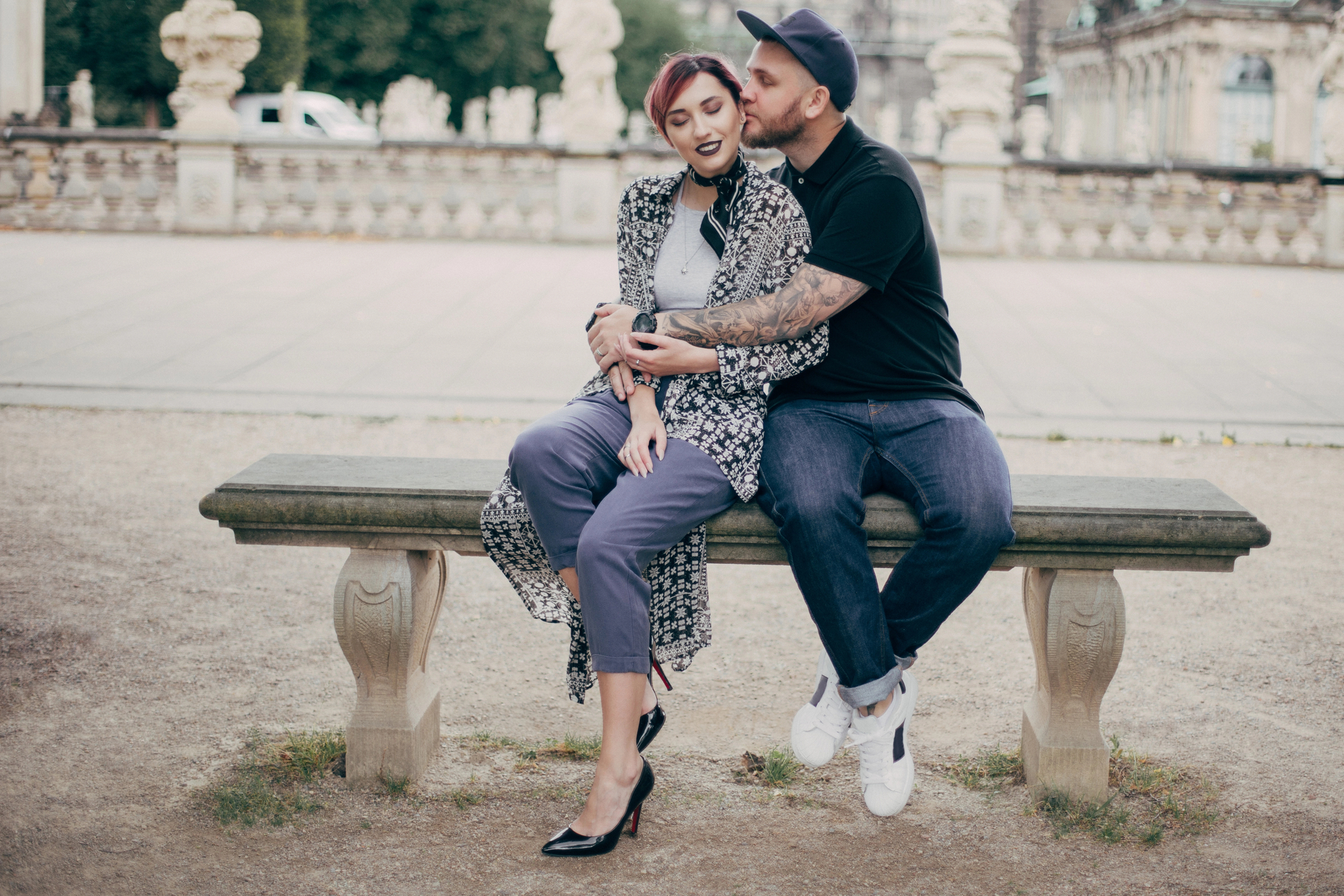 A couple sits on a stone bench outdoors. The man, wearing a cap and dark clothing, embraces the woman from behind, smiling. The woman, wearing a patterned jacket and purple pants, closes her eyes and smiles. The background features historical architecture and statues.