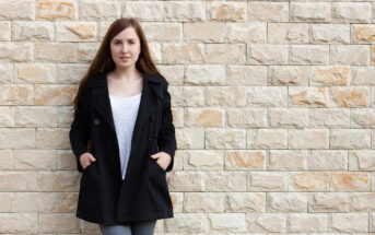 A person with long brown hair stands against a beige brick wall. They are wearing a black coat over a white shirt and have their hands in their coat pockets. The person looks directly at the camera with a neutral expression.