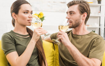 A woman and a man are sitting on a yellow couch, both wearing green shirts, and enjoying glasses of white wine. They are facing each other, smiling, and appear to be in a relaxed and comfortable setting with a bright, modern background.