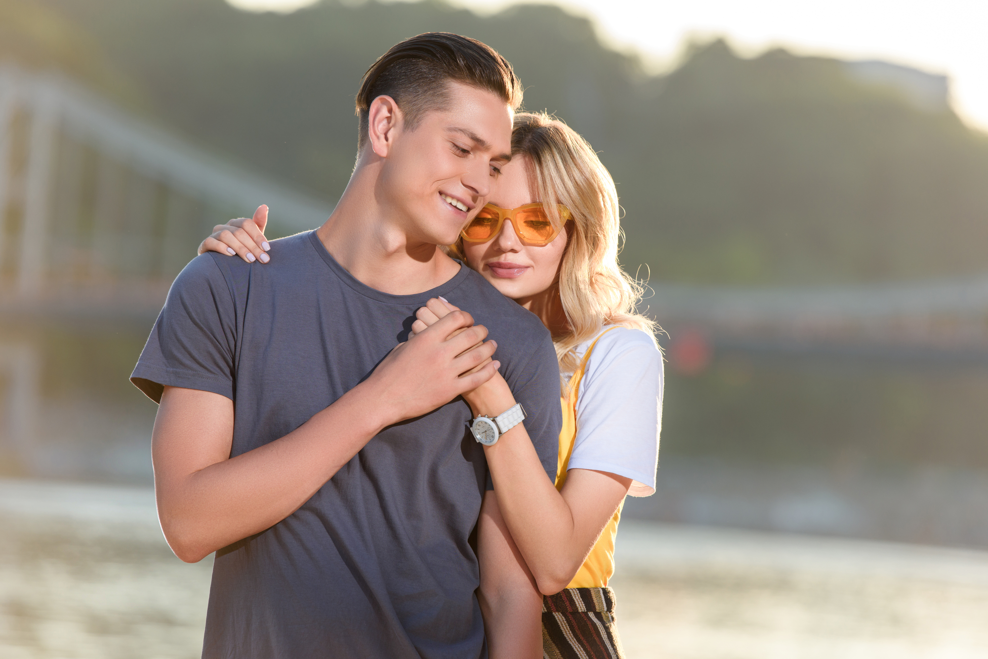 A smiling couple stands close together outdoors by the water. The man wears a blue t-shirt and the woman, who wears orange sunglasses and a white t-shirt, rests her head on his shoulder while hugging him. A bridge and trees are visible in the background.