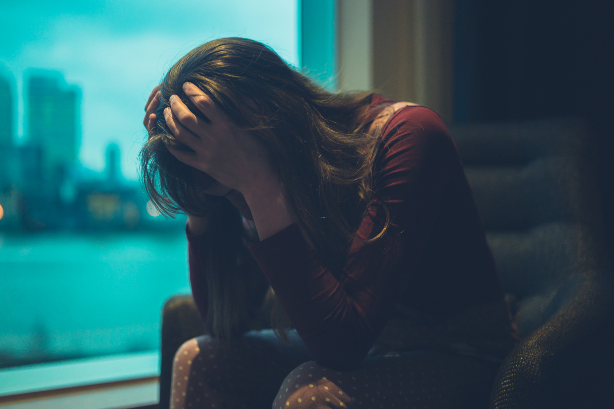 A person with long hair, dressed in a maroon top and patterned leggings, is seated and leaning forward with their hands holding their head, conveying a sense of distress or frustration. The background shows a window with a view of a city skyline and water.