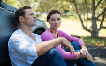 A man and a woman sit on a paved surface next to a car under the shade of trees. The man is wearing a white shirt and appears to be talking, while the woman in a pink top looks directly at the camera with a serious expression.