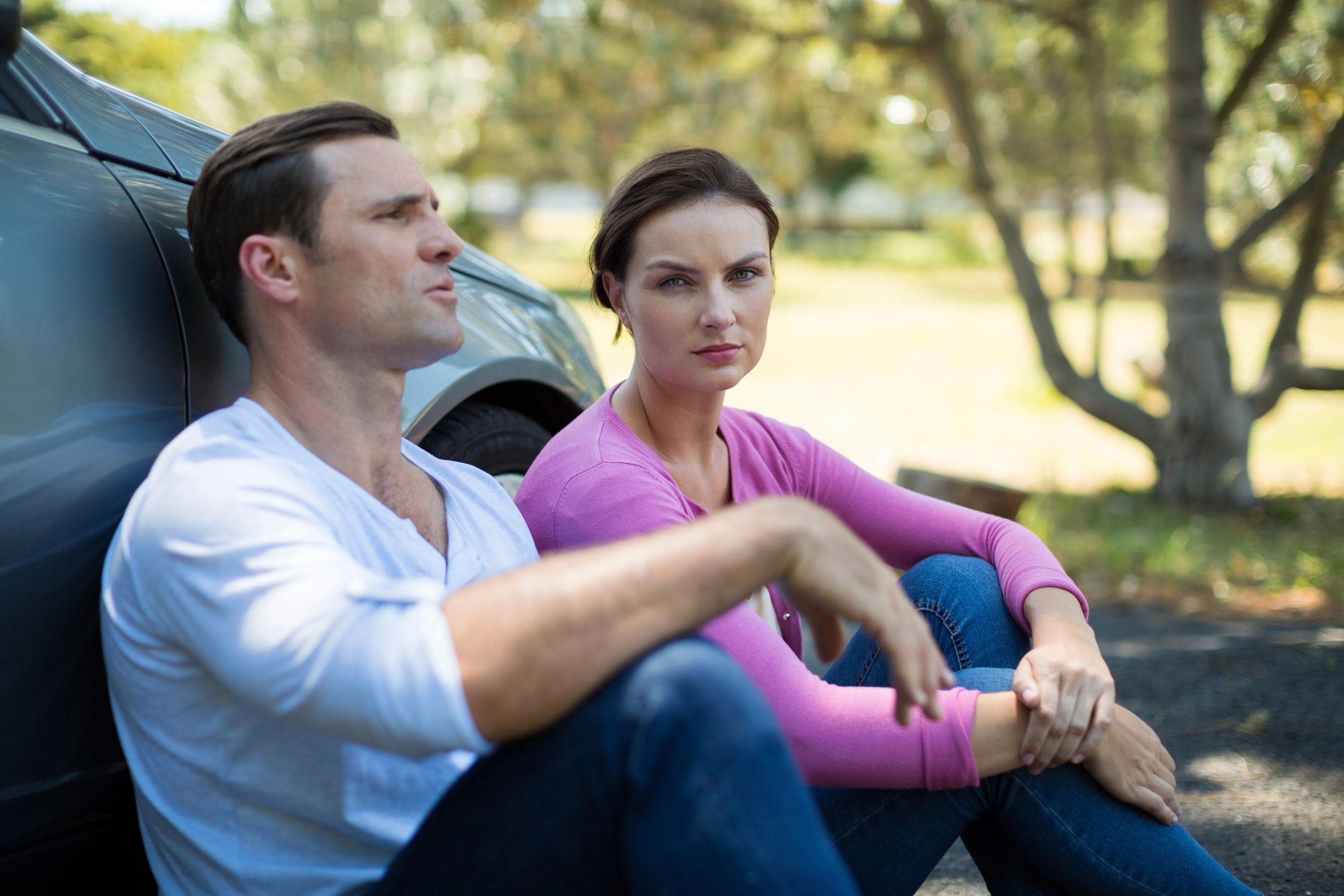 A man and a woman sit on a paved surface next to a car under the shade of trees. The man is wearing a white shirt and appears to be talking, while the woman in a pink top looks directly at the camera with a serious expression.