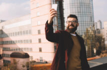 A bearded man wearing glasses and a burgundy jacket smiles while taking a selfie with his smartphone. He stands on a rooftop with pink and glass buildings in the background on a sunny day.