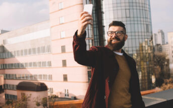 A bearded man wearing glasses and a burgundy jacket smiles while taking a selfie with his smartphone. He stands on a rooftop with pink and glass buildings in the background on a sunny day.