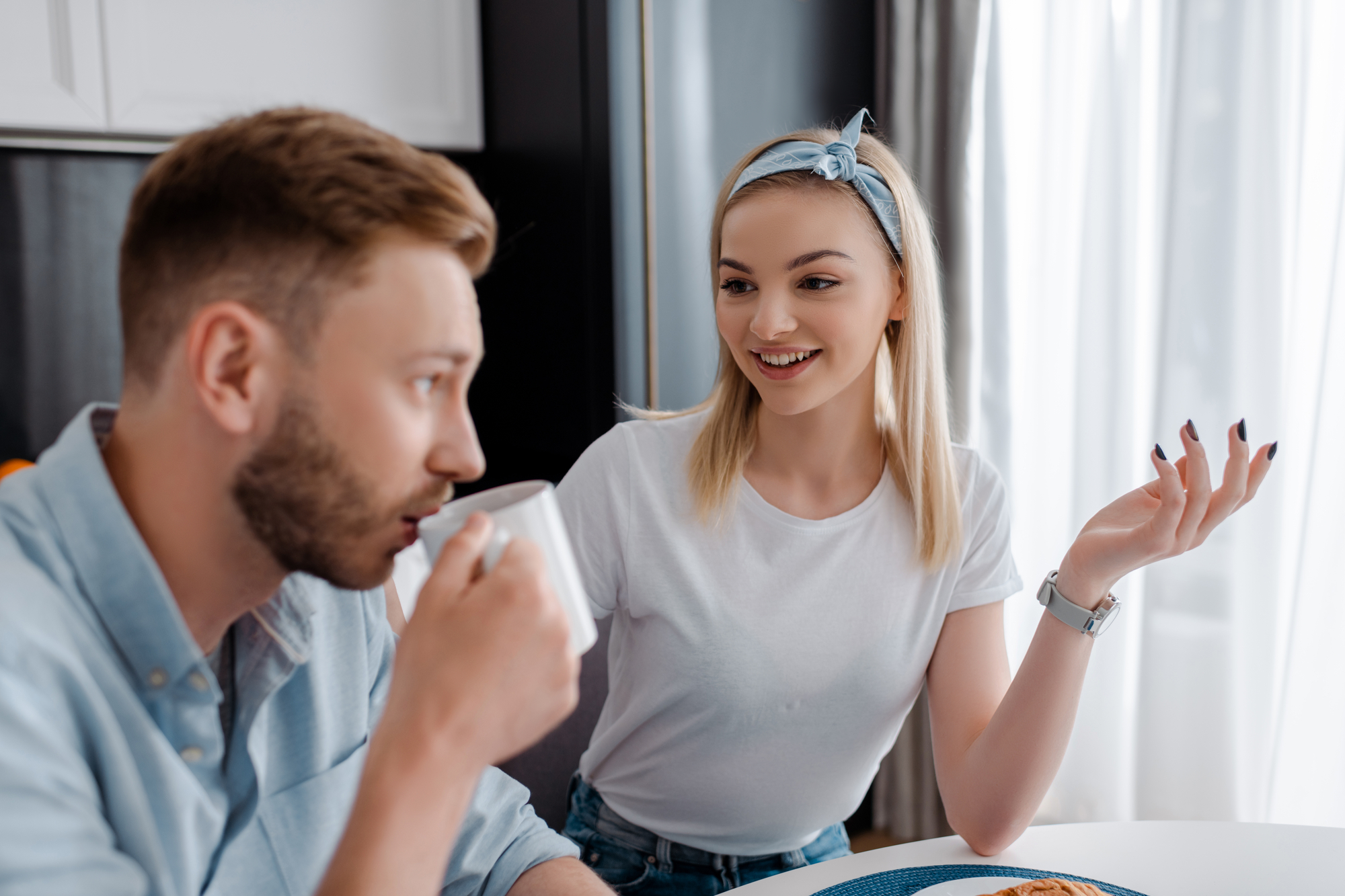 A woman with long blonde hair wearing a white shirt and a headband gestures while smiling at a seated man with short hair, a beard, and a blue shirt. The man is sipping from a white mug. They appear to be sitting at a dining table in a bright room.