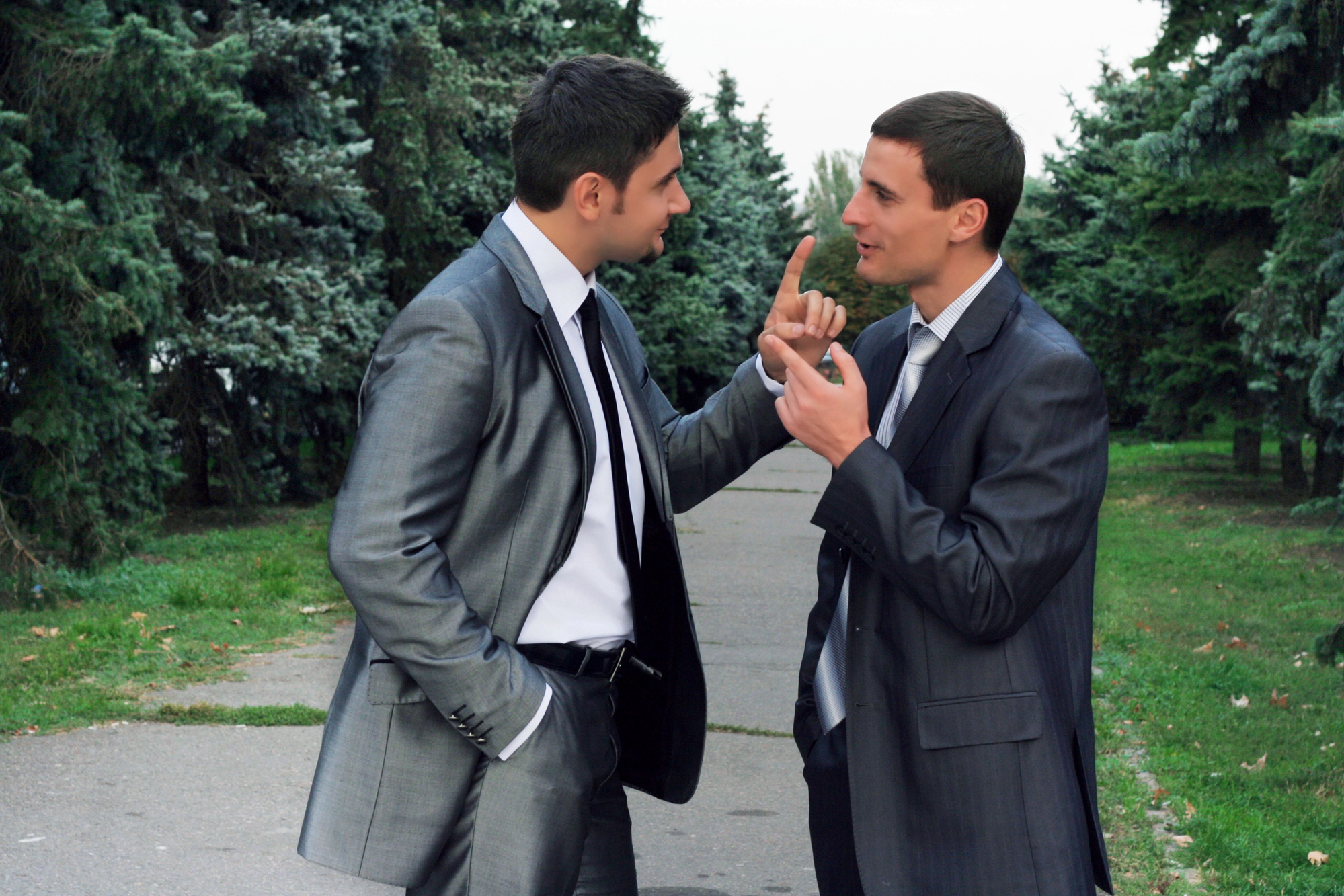 Two men in suits stand on a path lined with trees, engaged in an animated conversation. One man gestures with his hand, raising his index finger, while the other has his hand in his pocket and appears to be listening intently. They are surrounded by greenery.