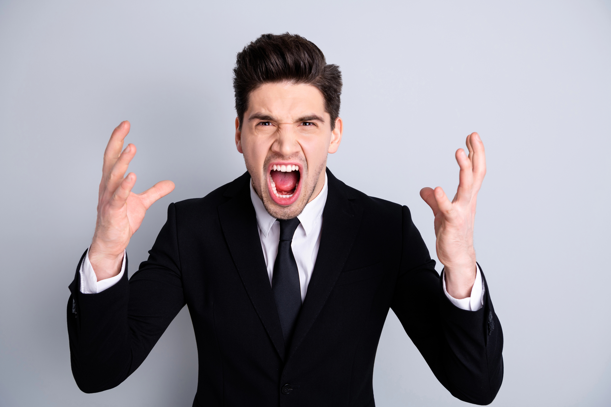 A man in a black suit and tie stands against a plain, light gray background. He appears to be yelling with his mouth wide open, eyebrows raised, and arms raised with fingers spread out, conveying a strong emotion, possibly frustration or anger.
