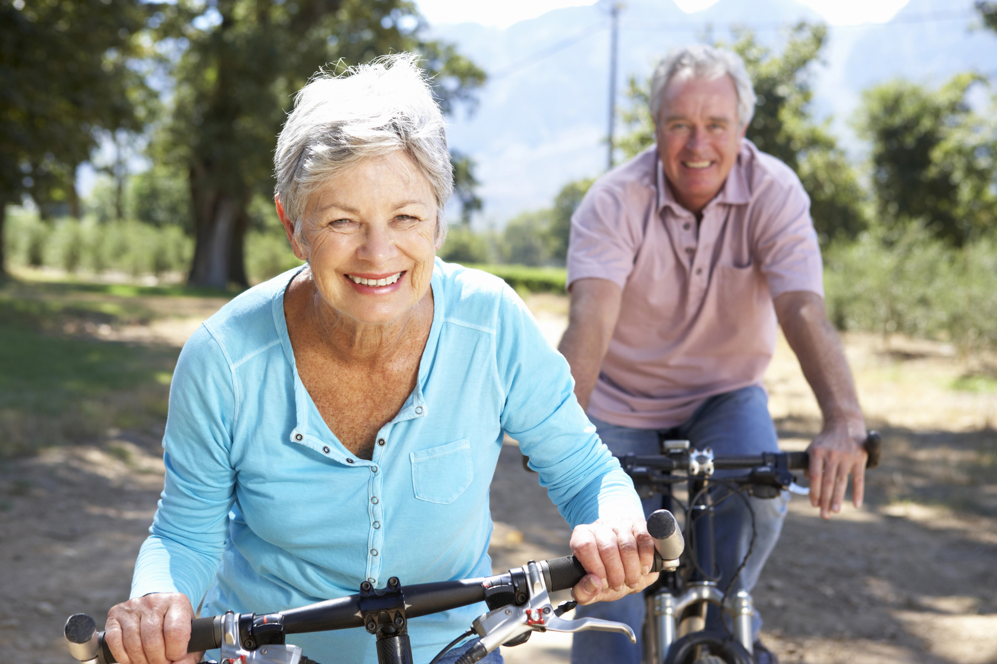 A happy elderly woman in a light blue top and an elderly man in a polo shirt ride bicycles on a sunny day. They are outdoors on a path surrounded by trees and greenery, smiling and enjoying their ride.