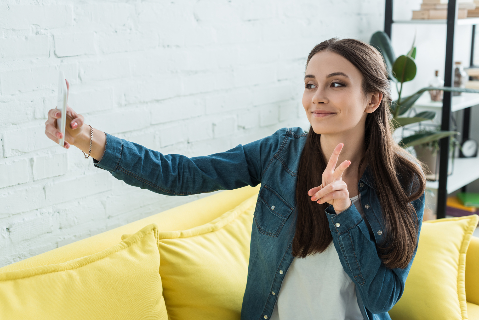 A young woman sits on a yellow couch, taking a selfie with her smartphone. She smiles and makes a peace sign with one hand. She wears a blue denim shirt and has long brown hair. The background features a white brick wall and some greenery.