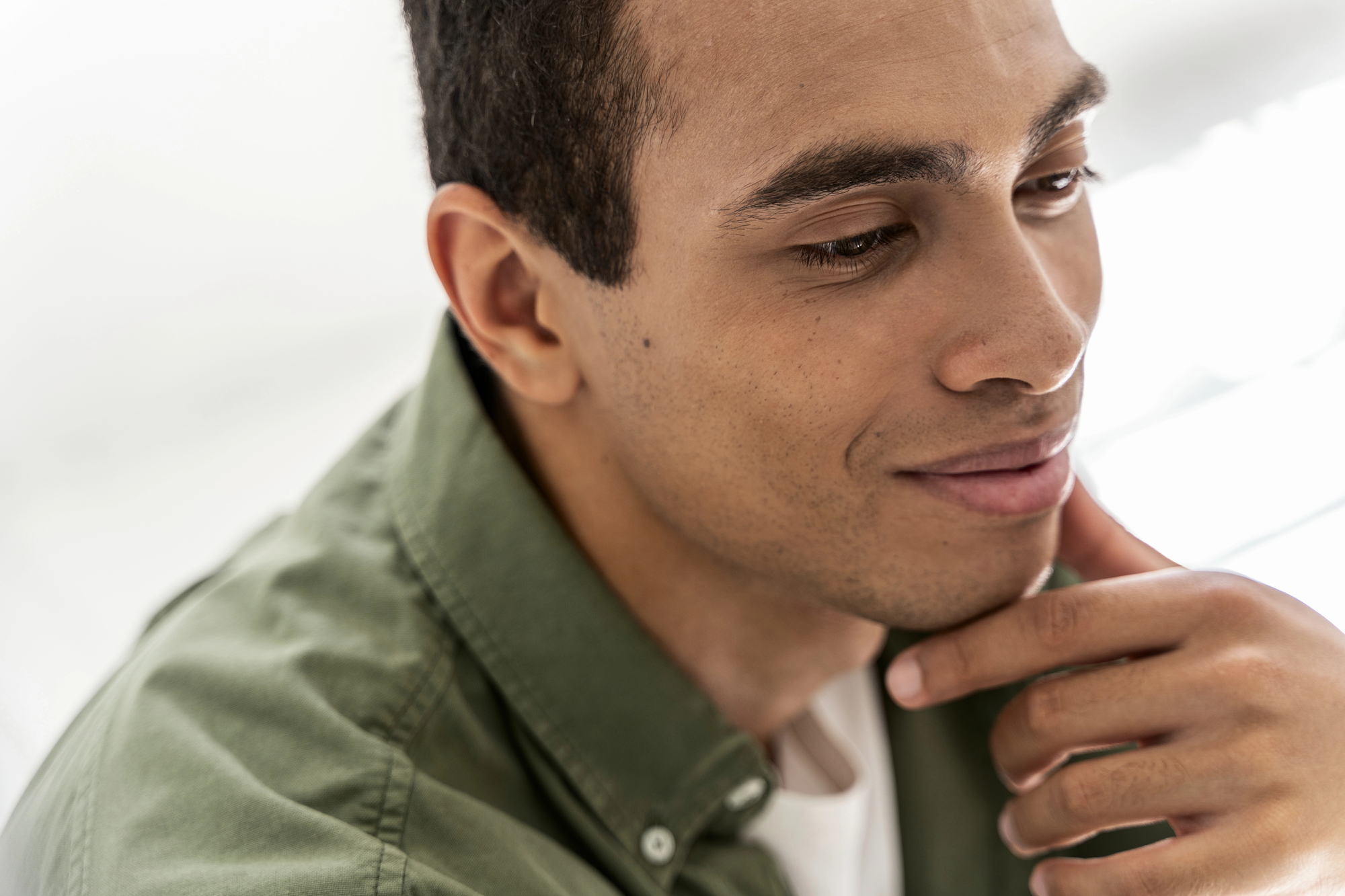 A young man with short dark hair is looking down thoughtfully, resting his chin on his hand. He is wearing a green collared shirt and appears to be indoors with soft, natural light illuminating his face.