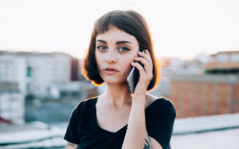 A woman with a short, dark bob haircut and tattoos on her arms holds a smartphone to her ear. She is wearing a black t-shirt and standing outdoors with blurred buildings in the background, likely at sunset, as the light is soft and warm.