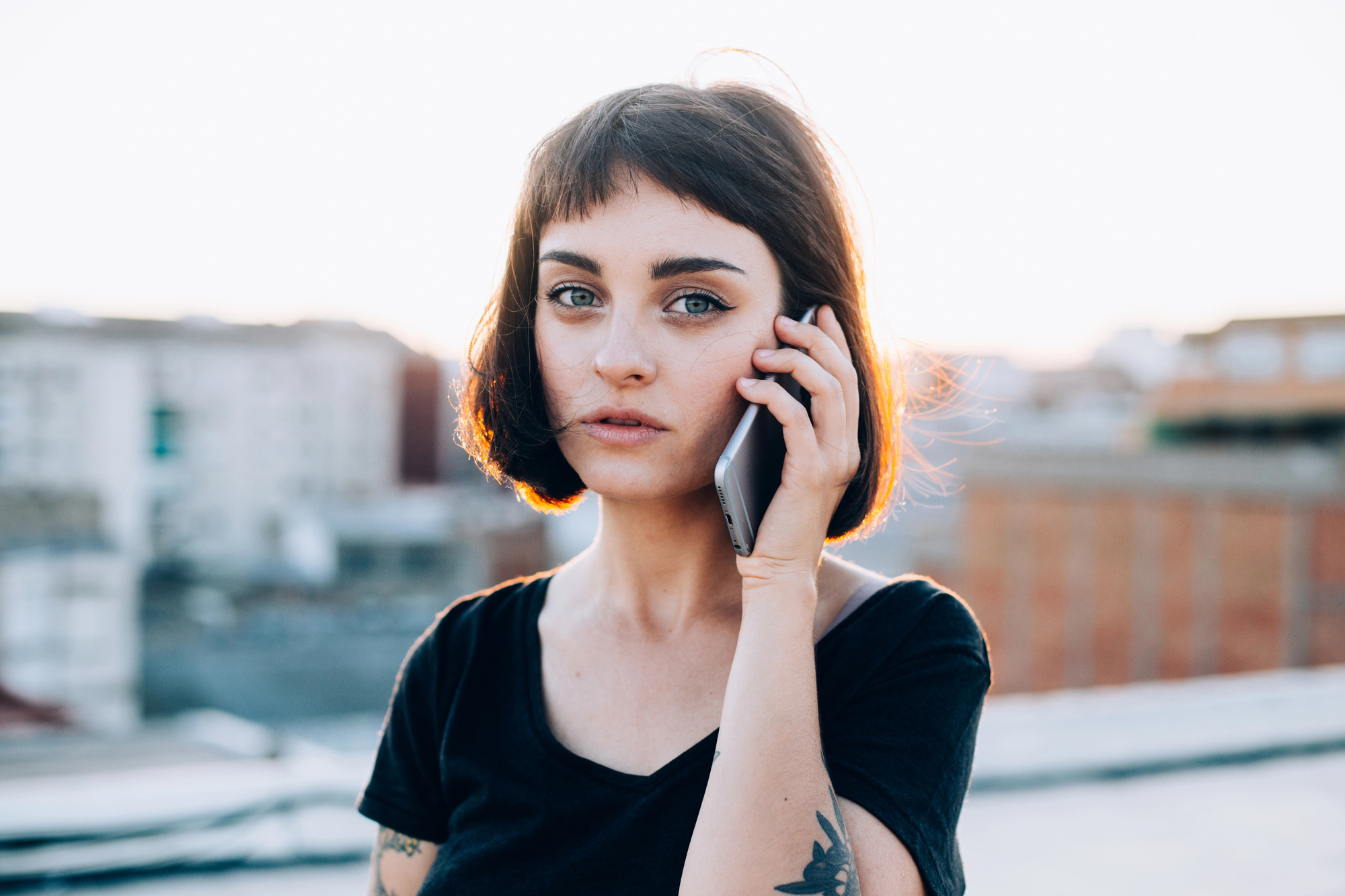 A woman with a short, dark bob haircut and tattoos on her arms holds a smartphone to her ear. She is wearing a black t-shirt and standing outdoors with blurred buildings in the background, likely at sunset, as the light is soft and warm.