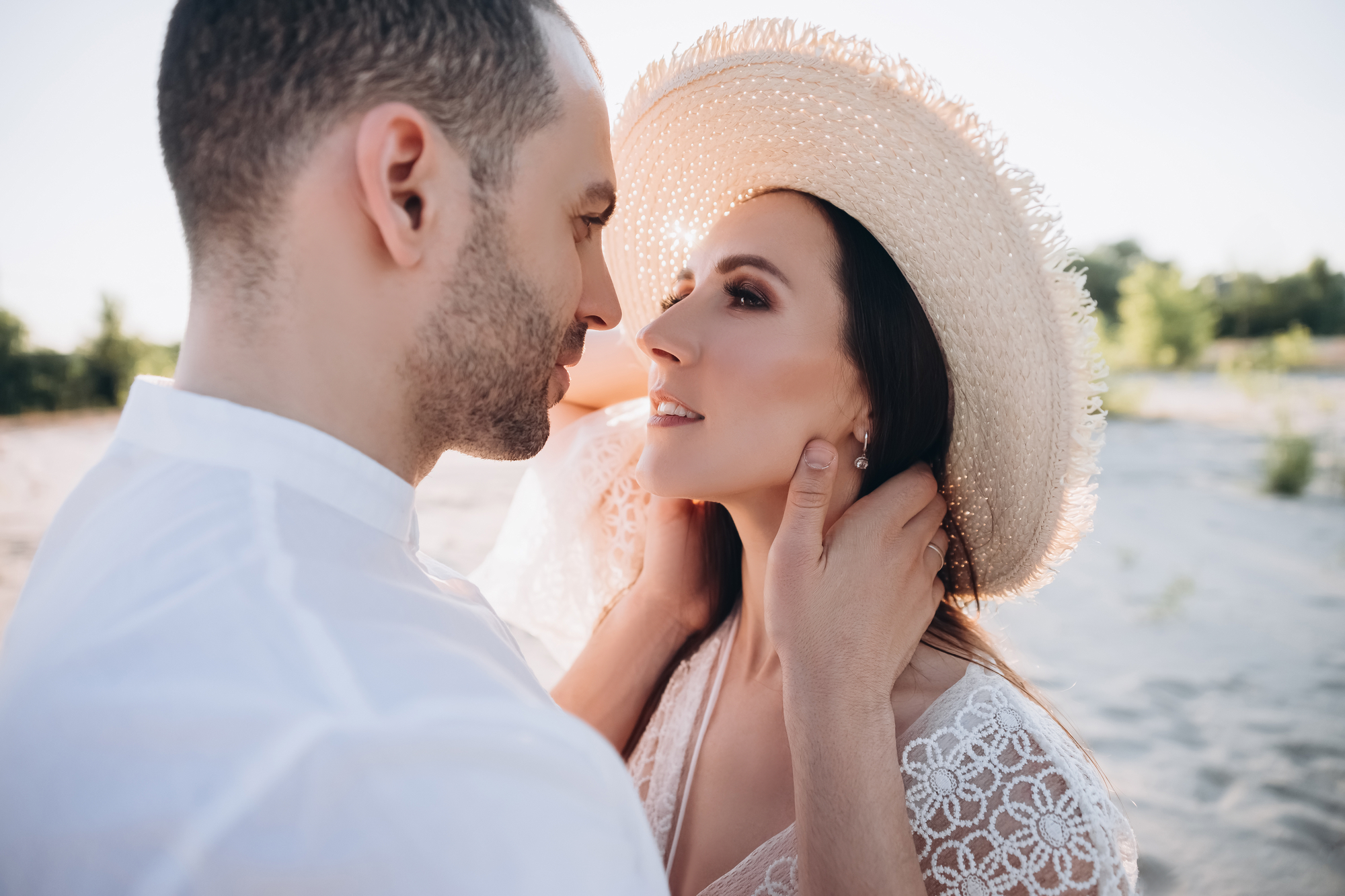 A man and woman share an intimate moment outdoors. The man, wearing a white shirt, gently holds the woman's face. The woman, in a lace dress and wide-brimmed straw hat, gazes up at him affectionately. The background features a sandy, sunlit landscape with greenery.