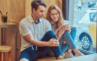 Two young adults sitting together by a window in a cafe, with a glass of green smoothie on a table nearby. They are both looking at a tablet and smiling. The young woman has curly hair and glasses, while the young man has short hair and is wearing a polo shirt.