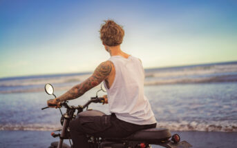 A person with tattoos on their left arm is sitting on a motorcycle at the beach. They are wearing a white tank top and black pants, looking towards the ocean as the sun begins to set. The sky is clear and blue.