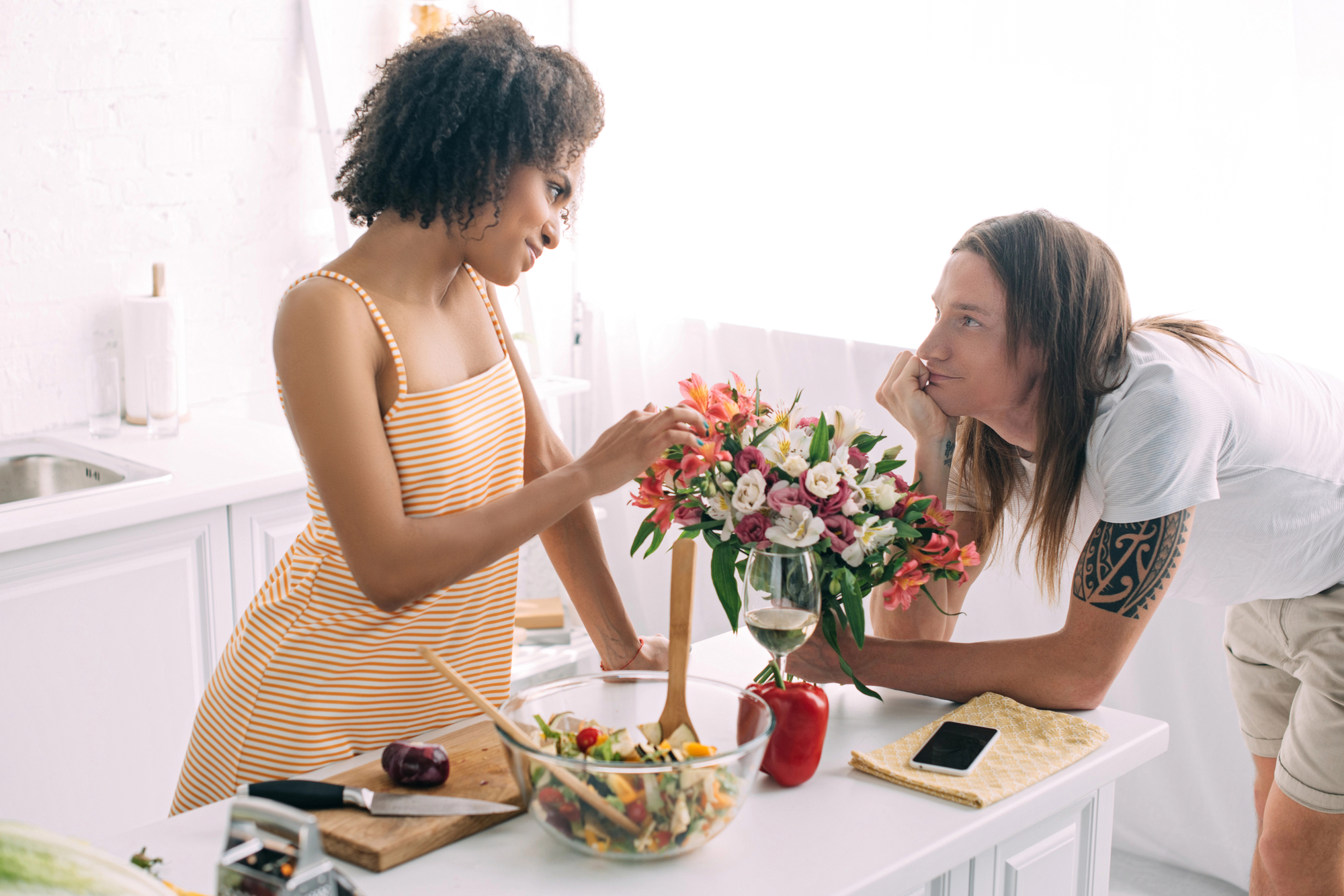 A woman in a striped dress arranges flowers in a kitchen while a man with long hair and a tattooed arm leans on the counter, watching her. A salad bowl, cutting board, mobile phone, and wine glass are on the counter. They both seem engaged in conversation.