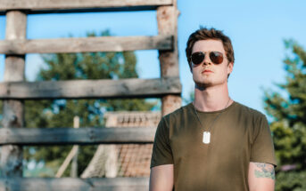 A man wearing sunglasses and a green t-shirt stands outdoors with a serious expression. He has a dog tag necklace and a tattoo on his left arm. The background features a rustic wooden structure and greenery under a clear blue sky.