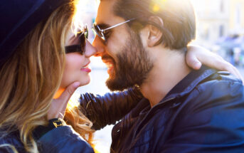 A couple dressed in black leather jackets, wearing sunglasses, and embracing each other closely while smiling. They are about to kiss, with sunlight softly illuminating them from behind, giving a warm and romantic atmosphere.