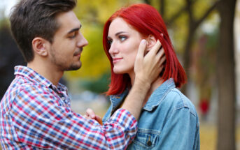 A young man with brown hair, wearing a plaid shirt, gently holds the face of a young woman with red hair, wearing a denim jacket. They are outdoors with trees in the background, looking into each other’s eyes with a serene expression.