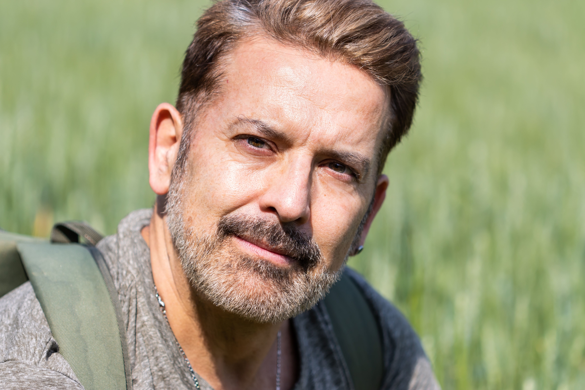 A man with short brown hair and a beard is outdoors in a green field, wearing a gray shirt and carrying a backpack. He has a calm expression and is looking directly at the camera.