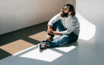 A man with a beard sits on the floor in a sunlit room, leaning against a wall. He is wearing a white shirt and blue jeans, with one hand resting on his knee and the other on the floor. Sunlight creates rectangular patterns on the floor and wall.