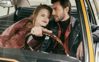 A couple is sitting in the front seat of a car, with the woman leaning on the man who is holding the steering wheel. Both are wearing leather jackets and smiling affectionately at each other, creating a warm, intimate moment.