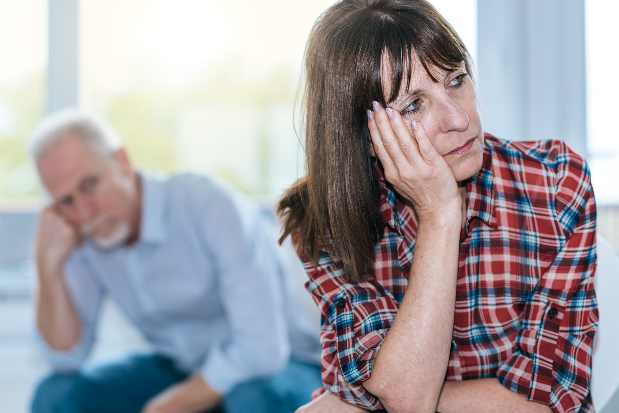 A woman in a plaid shirt rests her head on her hand and looks pensive. In the background, a man with a gray beard sits with his head resting on his hand, appearing equally contemplative. Both have expressions suggesting sadness or deep thought.