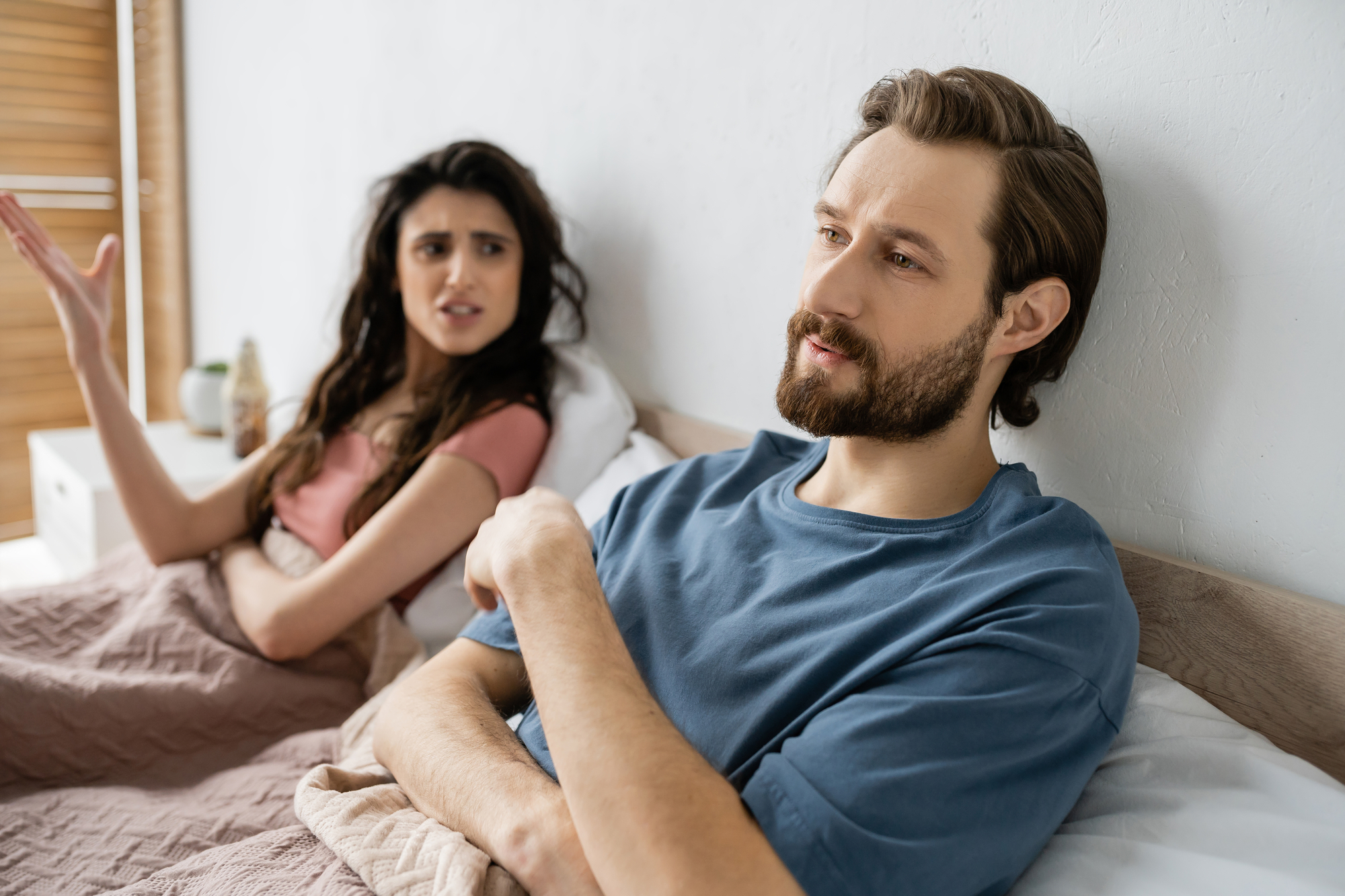 A man with a beard is sitting up in bed, wearing a blue shirt, and looking thoughtful while a woman with long dark hair, wearing a pink shirt, is gesturing expressively and looking at him with a concerned expression. They are both under a beige blanket.