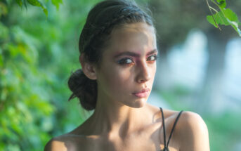 A woman with braided dark hair looks intently at the camera. She wears subtle makeup with a touch of blush and pink eyeshadow. She has a sleeveless top and is surrounded by green foliage, with sunlight casting gentle shadows on her face.