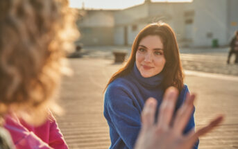 A young woman with long brown hair and wearing a blue sweater is smiling while listening attentively to an unseen person gesturing in the foreground. They are outdoors, with a calm, sunlit setting and buildings in the background.