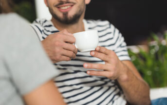 A person with a beard and mustache, wearing a white and black striped shirt, is smiling and holding a white cup with both hands. A portion of another person's shoulder is visible in the foreground. There are green plants in the background.