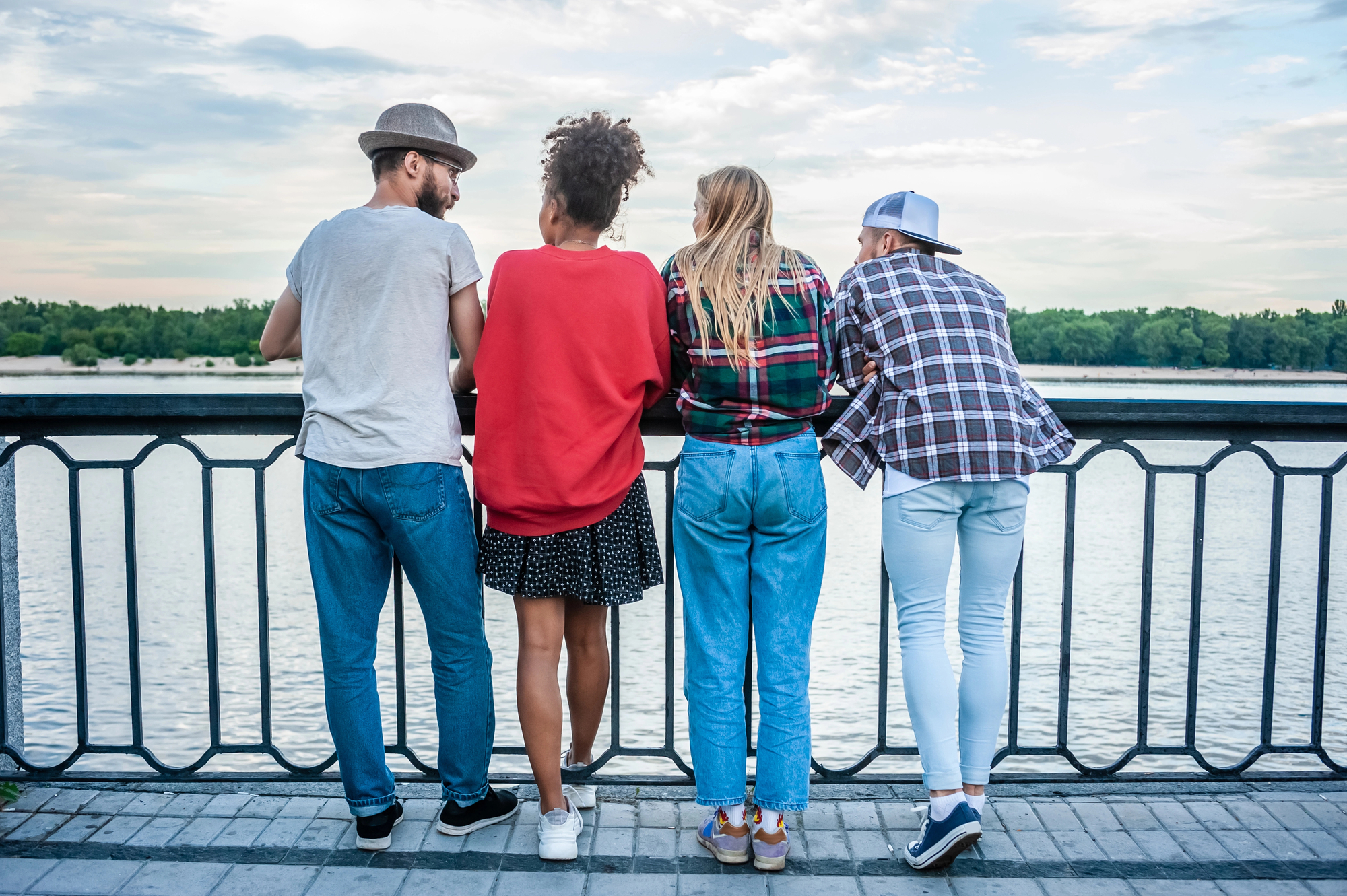 Four people are standing on a paved walkway, leaning over a black metal railing and looking out at a body of water. They are casually dressed, with one person wearing a hat and two wearing plaid shirts. Trees are visible in the background under a cloudy sky.