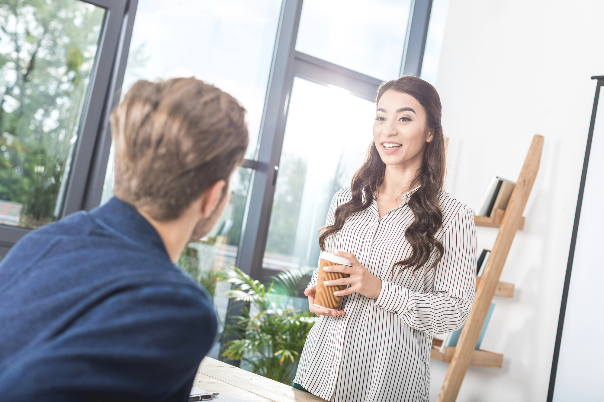 A woman with long hair, wearing a striped shirt, stands holding a cup and smiling while talking to a man sitting with his back to the camera in a modern office space with large windows and a wooden ladder shelf in the background.