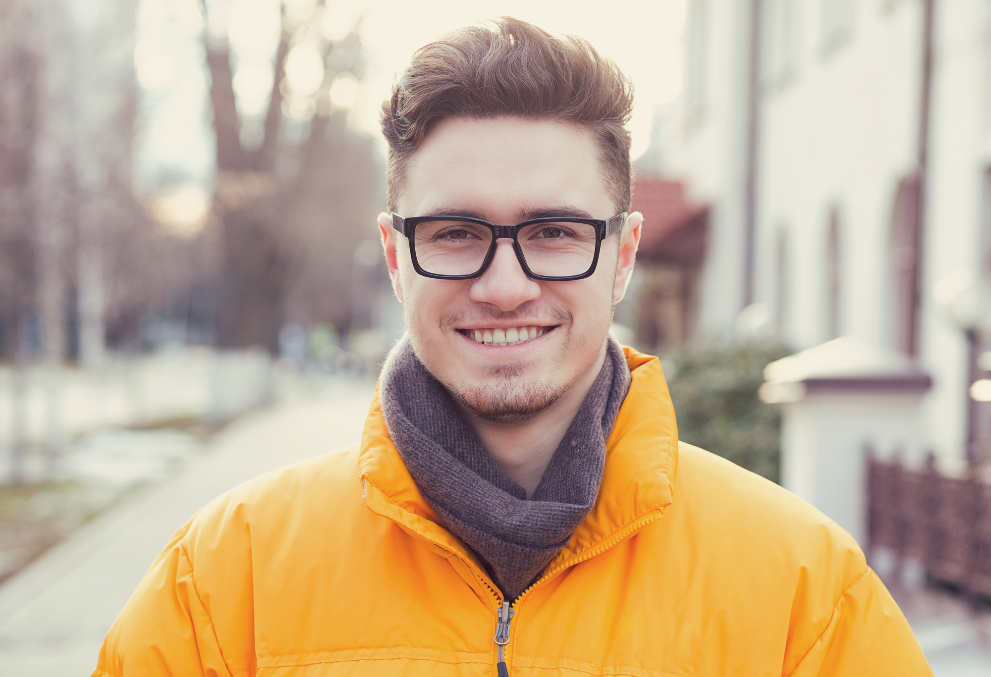 A person with short brown hair, black-framed glasses, and a slight beard and mustache smiles at the camera while wearing an orange jacket and a gray scarf. The background features a blurred street scene with trees and buildings.