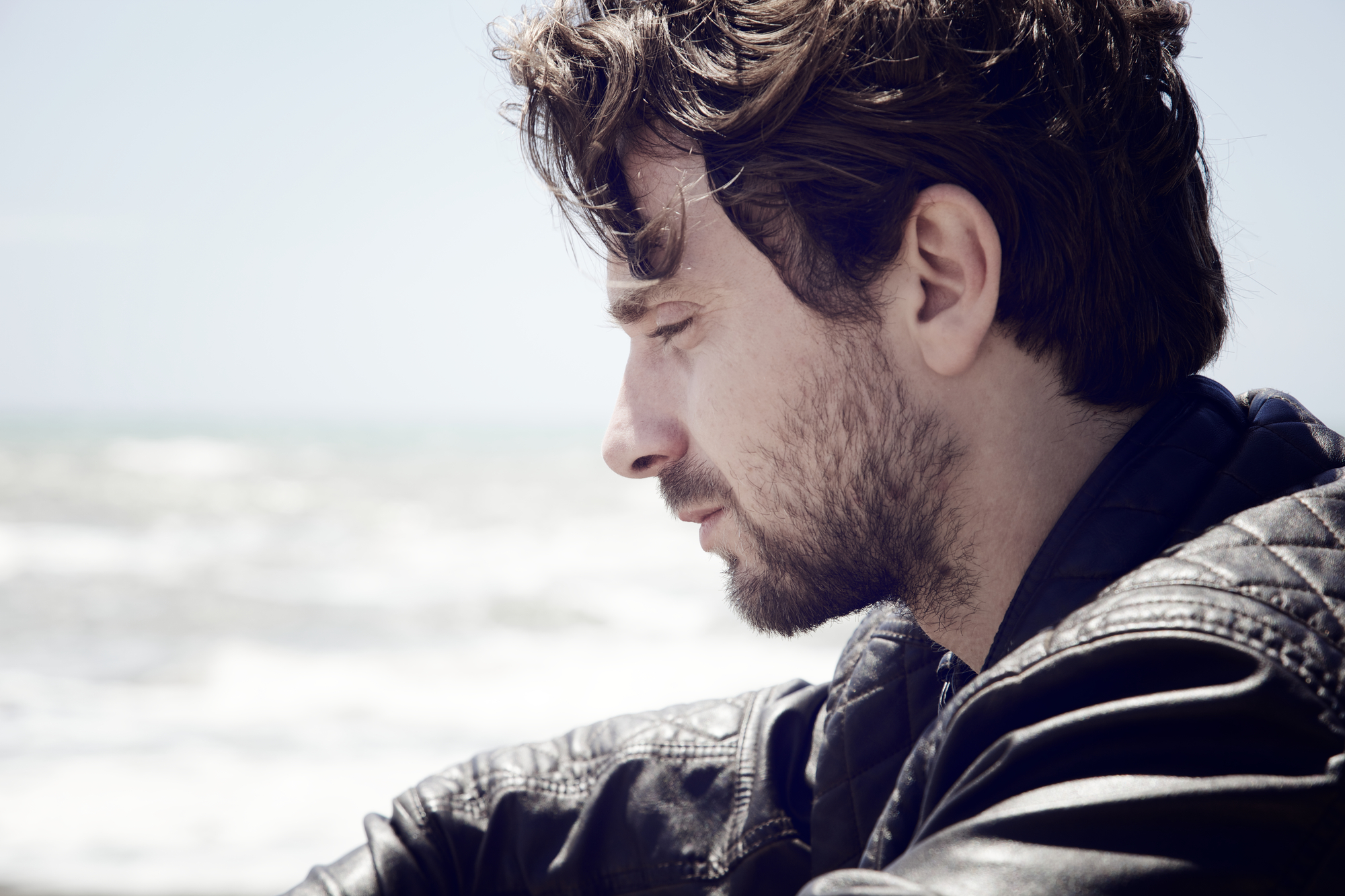 Side view of a young man with wavy brown hair and a beard wearing a black leather jacket. He appears deep in thought while looking down, with a blurred ocean background under a clear sky.